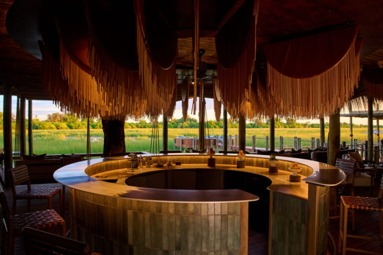 Circular outdoor bar with tiled counter under a thatched roof, overlooking a green marsh landscape at sunset/light.”] ,