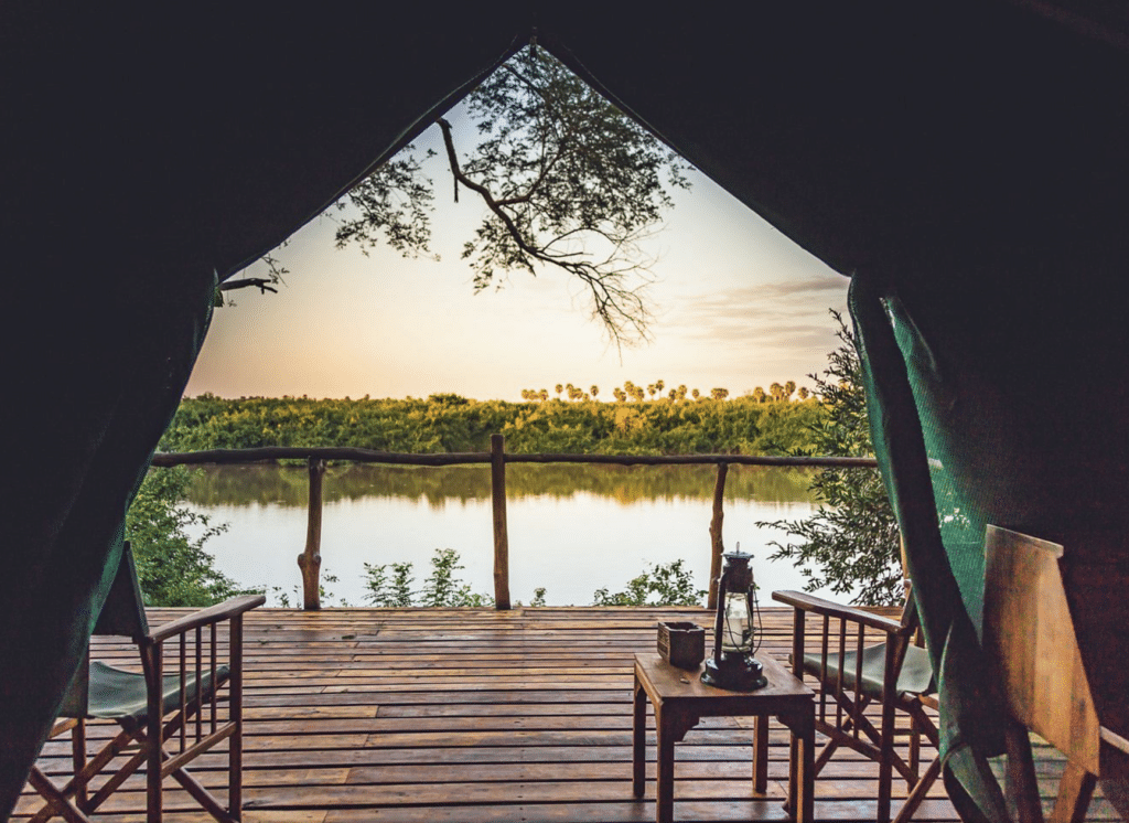 Open tent doorway revealing a wooden deck facing a calm lake and forest at sunset, with chairs and a lantern nearby?