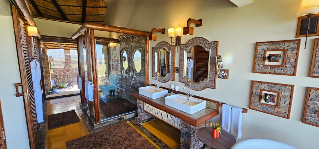Luxurious bathroom with twin rectangular sinks, stone-framed mirrors, wooden beams, and a glass-enclosed shower; warm lighting and an outdoor landscape visible through the door.