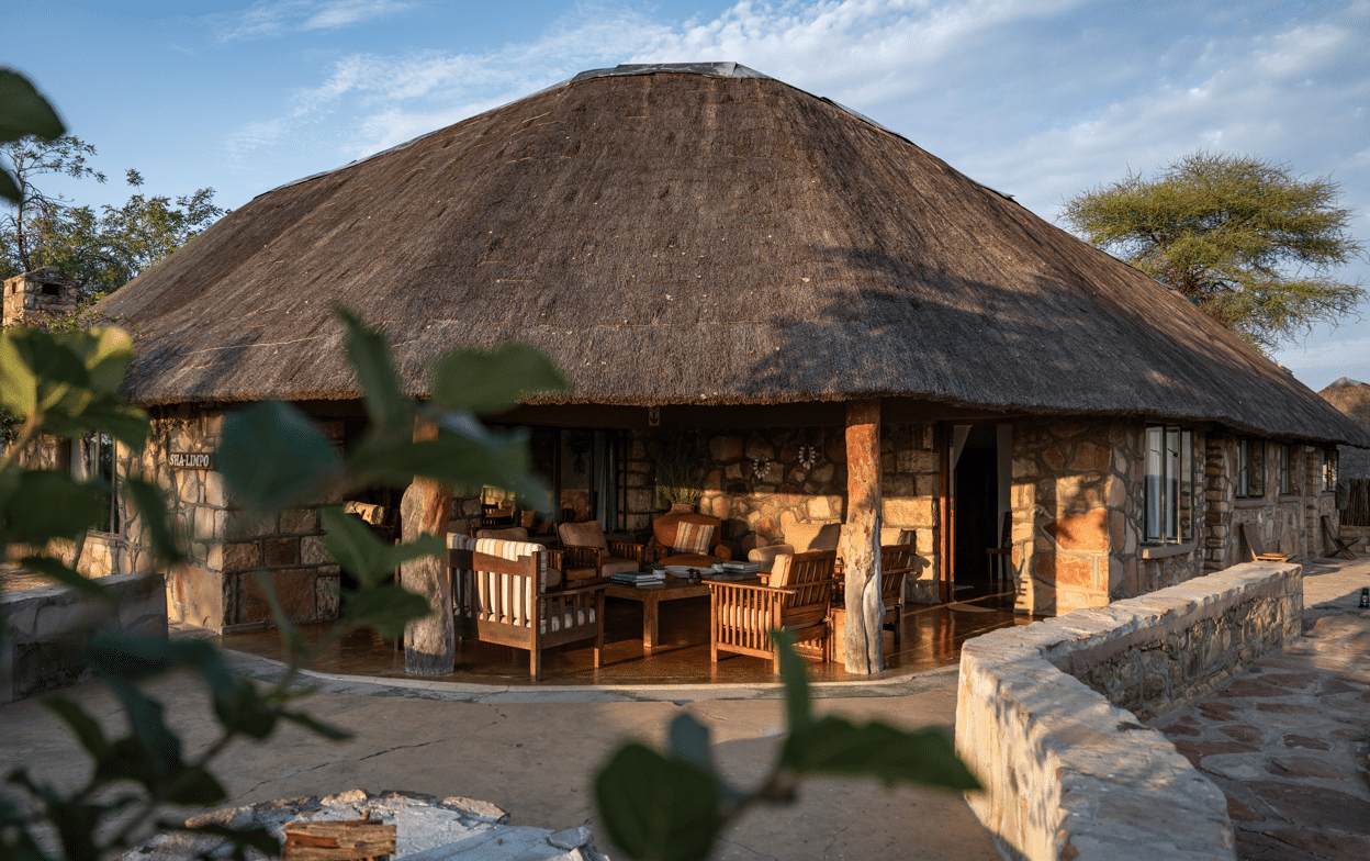 Thatched-roof circular cottage with a stone exterior and an open veranda furnished with wooden chairs and a coffee table.