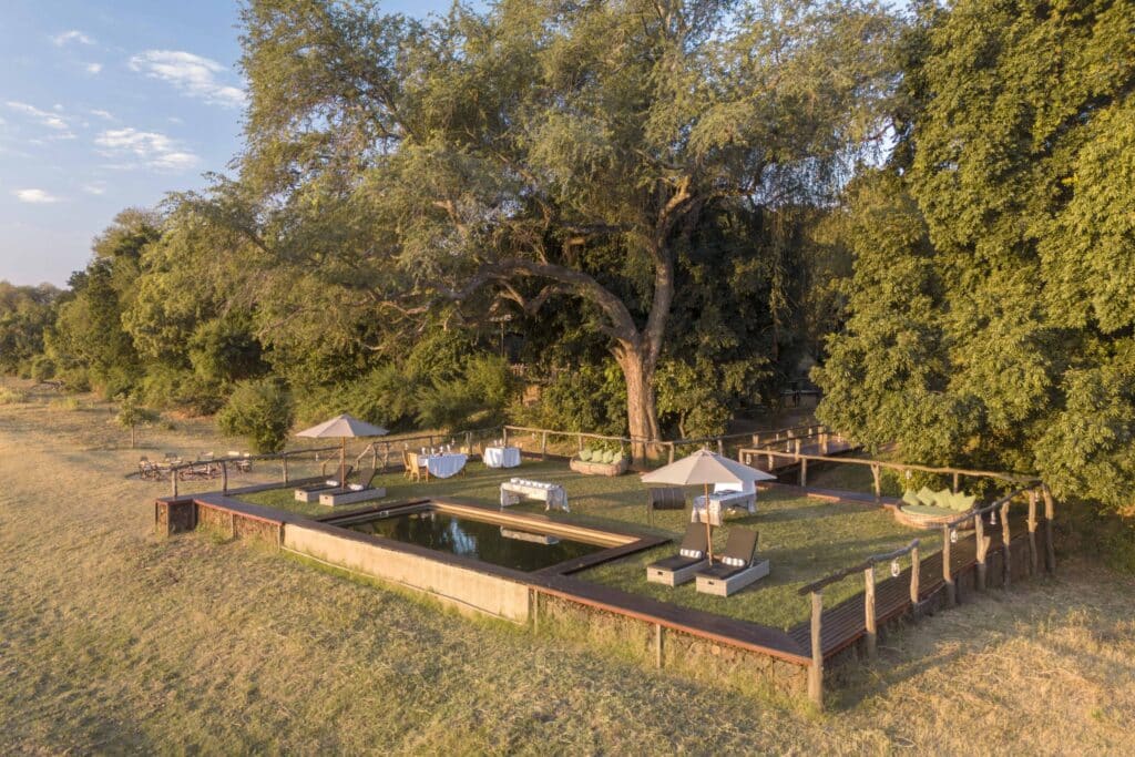 Outdoor lounge area under a large tree with tables, umbrellas, and cushioned seating by a low reflecting pool or water feature, fenced yard.