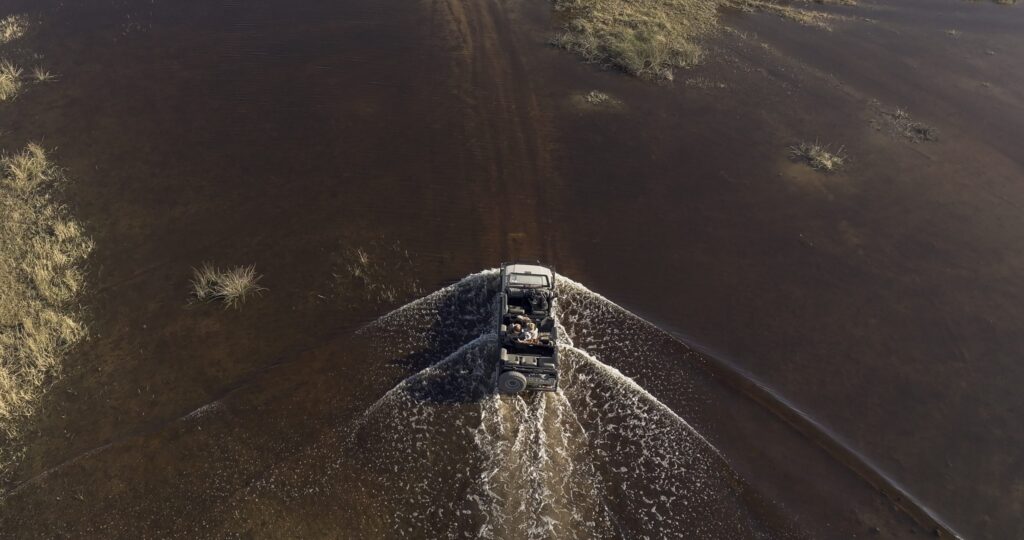 Aerial drone view of an off-road SUV crossing a shallow, muddy river, creating a foamy wake behind it.