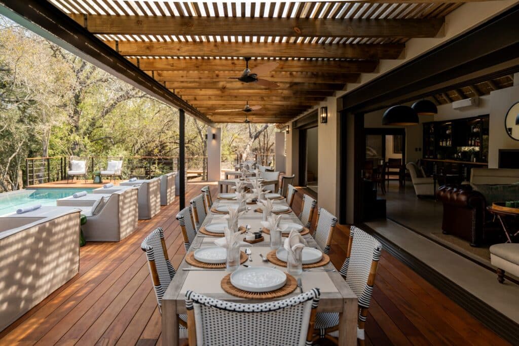 Outdoor covered dining area on a wooden deck, long table set with plates and napkins, beside a pool and lounge chairs.