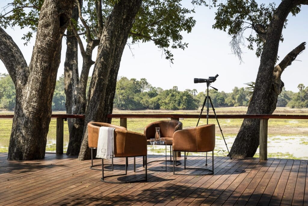 Outdoor wooden deck seating area framed by large trees; a telescope on a tripod stands behind the chairs.