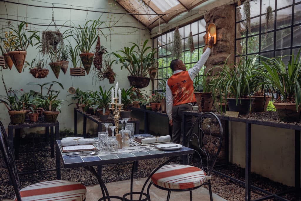 Restaurant table set for guests in a glasshouse with hanging plants and greenery; a staff member in an orange vest adjusts a wall light near stone wall.
