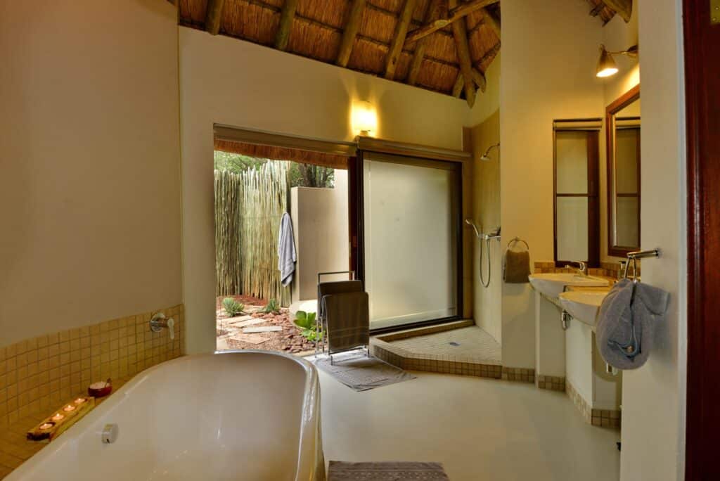 Bathroom with a white bathtub along the left, beige tiled edge, and a doorway opening to a courtyard with a bamboo fence.