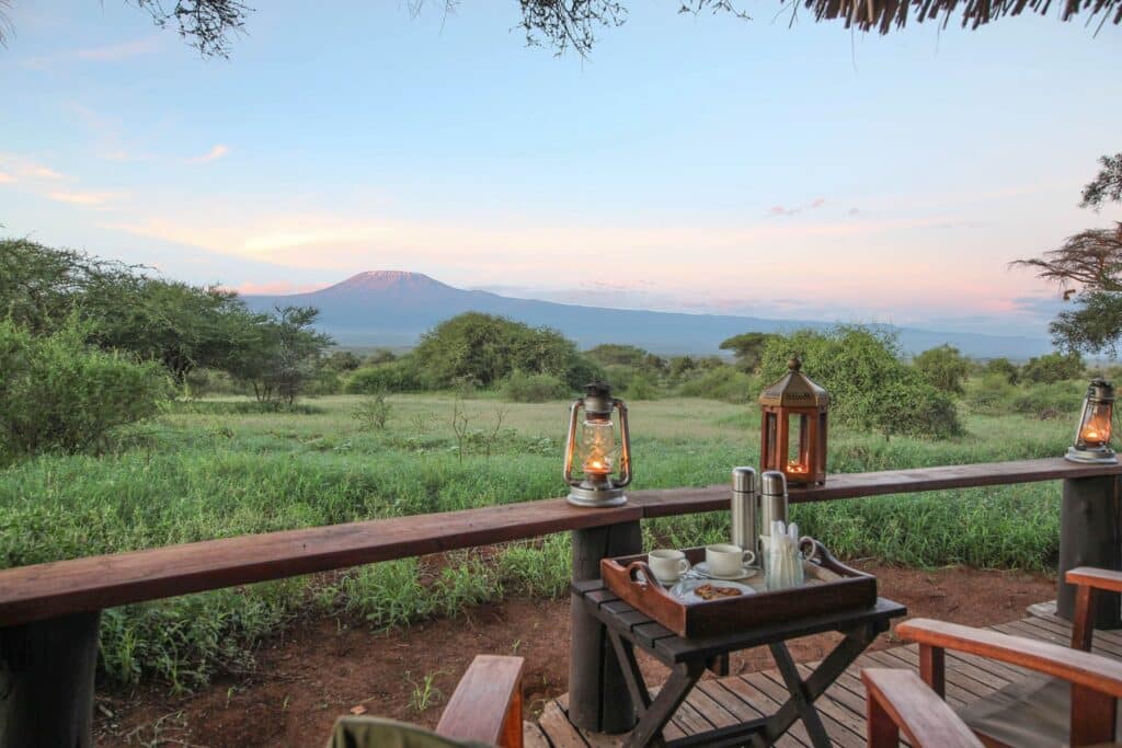 Deck with a tea service and glowing lanterns, overlooking a green savanna and a distant snow-capped mountain at dusk.