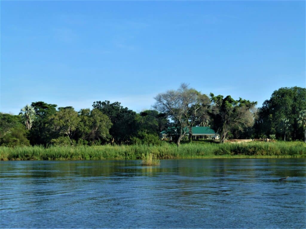 Riverside scene with calm water in the foreground, grassy bank, and a house with a green roof nestled among trees under a clear blue sky.