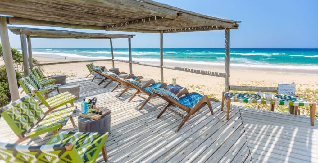 Wooden beach deck with lime-green patterned chairs facing turquoise ocean and white-sand beach under a sunny sky.