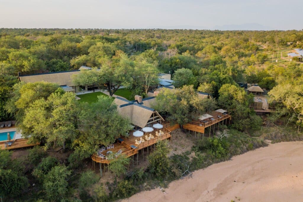 Aerial view of a forested resort with thatched-roof buildings and multiple wooden decks with outdoor seating nestled among trees near a sandy beach.