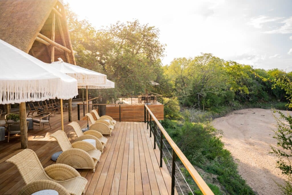 Wooden deck with wicker chairs under white thatched umbrellas, overlooking trees and a sandy path.