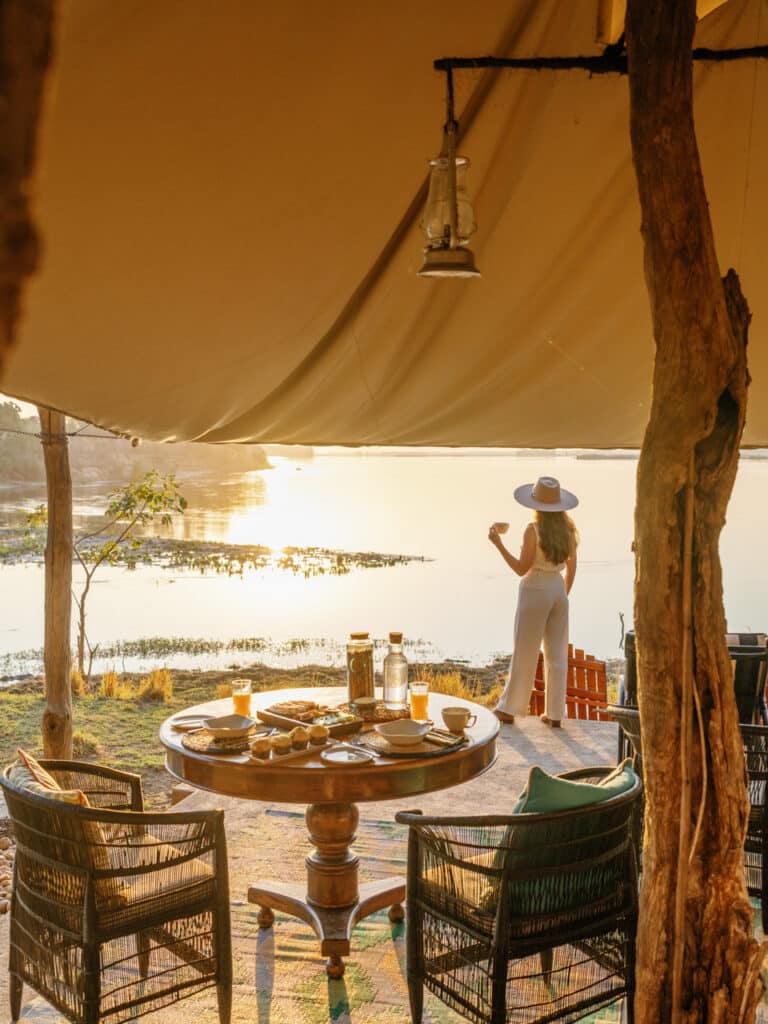 Sunset lakeside breakfast setup: a round table with pastries and drinks under a canopy, as a woman in white pants and a wide-brim hat gazes at the water with a glass in hand.