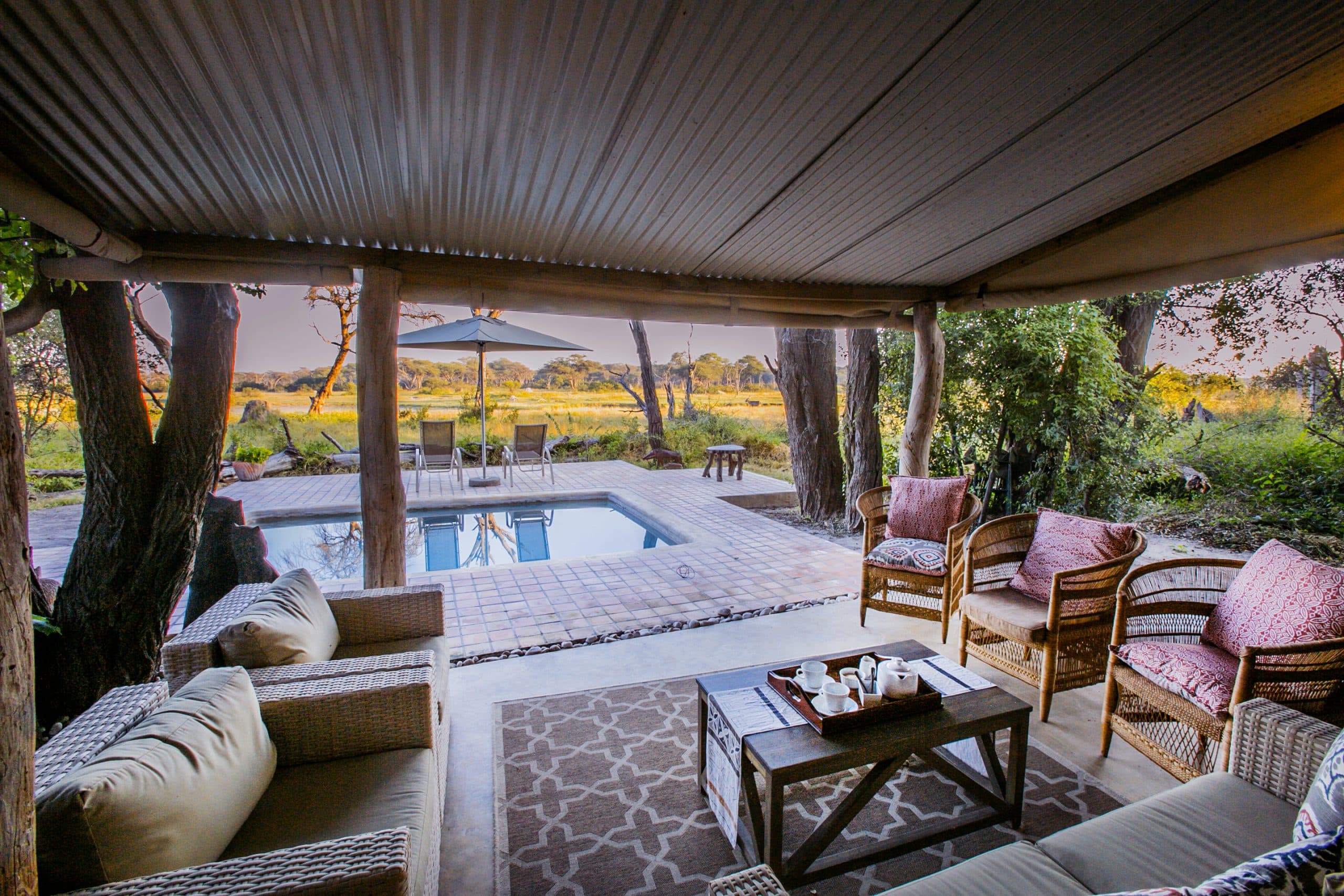 Outdoor covered patio with wicker sofas and a coffee table around a pool, overlooking a grassy savanna with trees in the distance.