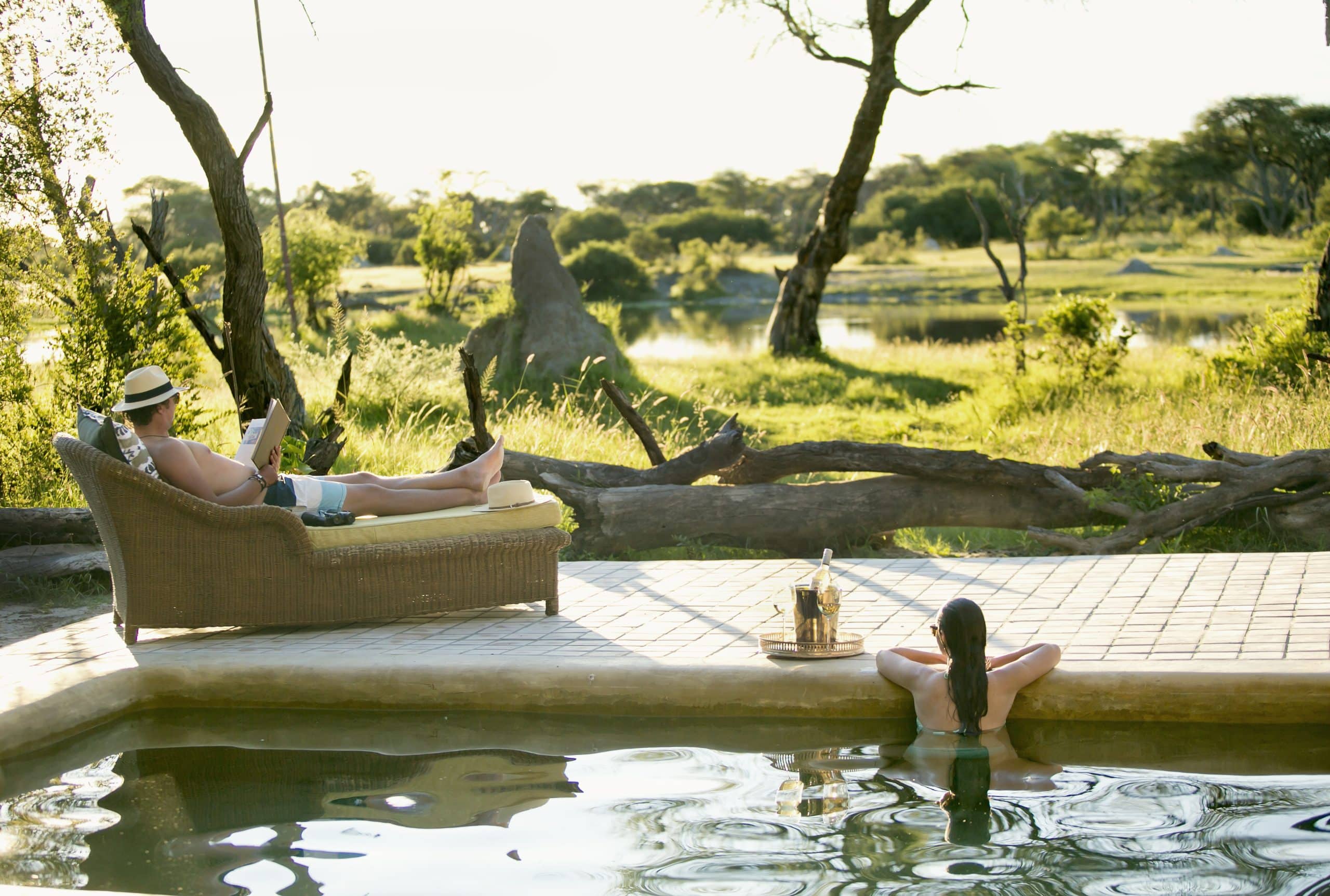Person in a sunhat lounging on a wicker chair beside a pool, reading a book in a sunny garden setting