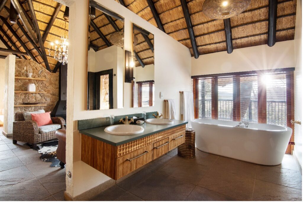 Rustic bathroom with a double-sink vanity, wood cabinets, and a freestanding white tub by large windows.
