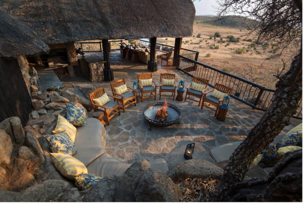 Outdoor stone patio with a circular fire pit surrounded by wooden chairs and cushions, overlooking a savannah landscape at dusk.