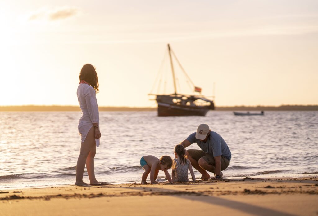 Family groups on a sandy beach at sunset, children playing in the shallow water while adults supervise; a sailboat anchored in the background.