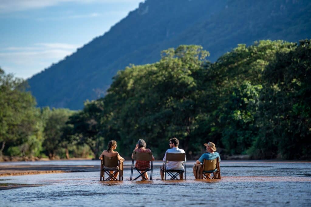Group of five people sitting in folding chairs at the water's edge, facing a lake with a forested hillside in the background.
