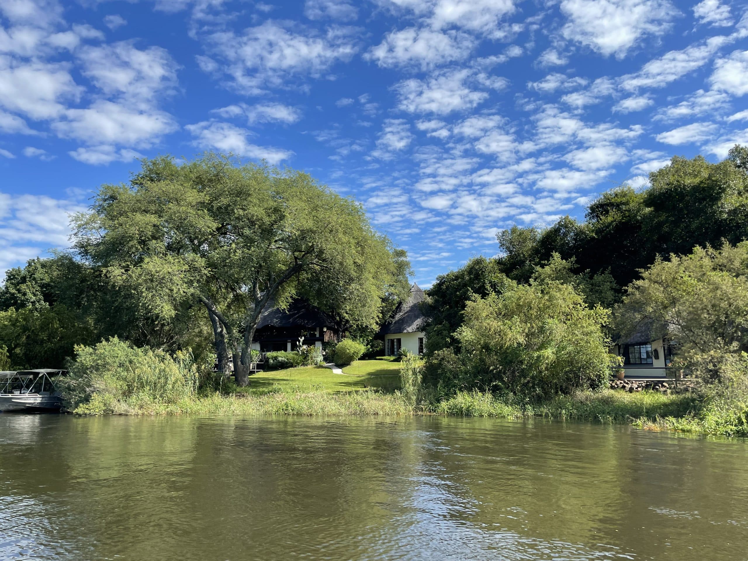 Calm riverfront with lush green trees along the bank and a thatched-roof house hidden among the foliage, under a bright blue sky with scattered clouds, left boat docked by the water.