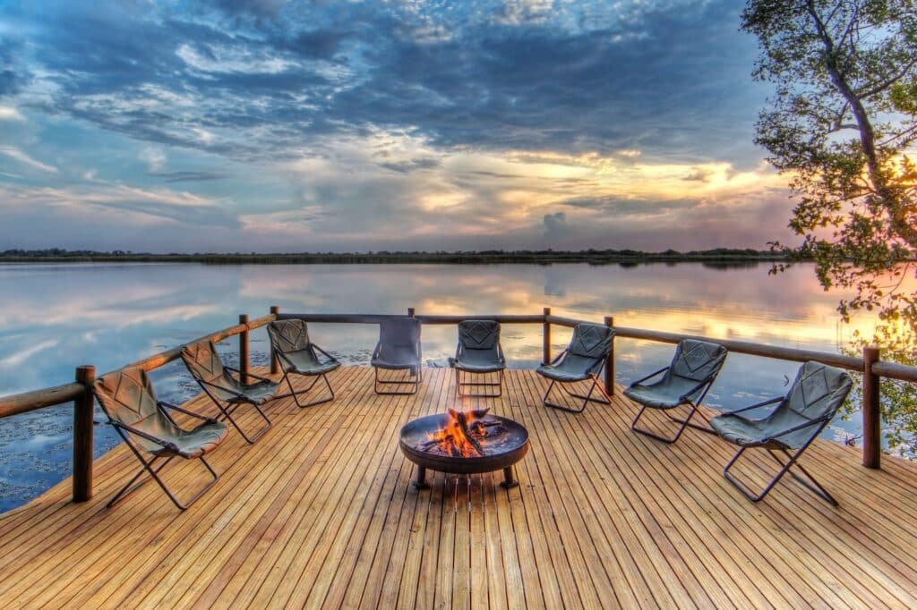 Wooden deck with cushioned chairs arranged around a fire pit overlooking a calm lake at sunset. A tree frames the right edge.