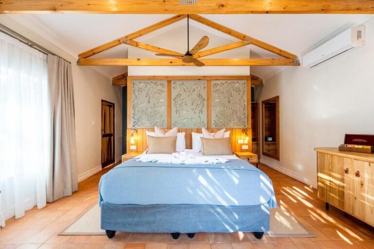 Bright hotel bedroom with a large blue bed, exposed wooden beams, and sunlit windows on the left.