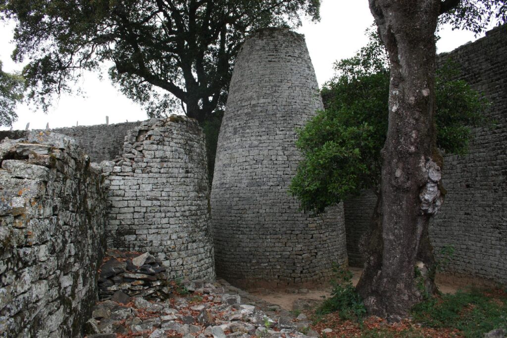 Ruined stone walls with a rounded tower surrounded by trees at a historic site.