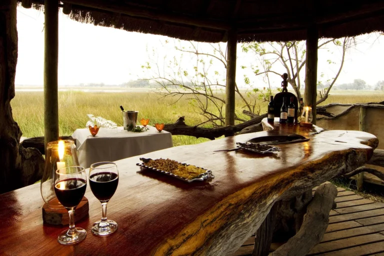 Open-air wooden bar built from a thick log under a thatched shelter, with wine glasses, candles, and a tray of snacks overlooking a marsh.