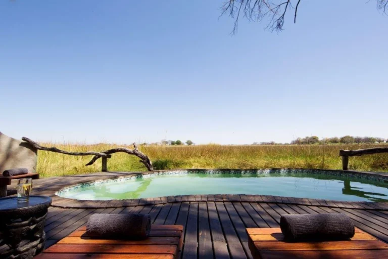 Outdoor wooden deck with a small pool, rolled towels on benches, and a branch railing overlooking tall grass under a clear blue sky.