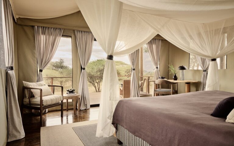 Luxurious safari tent bedroom with a canopy bed, sheer drapes, and a seating area overlooking an outdoor deck and trees.