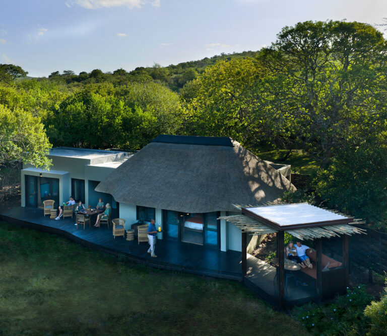 A modern villa with a large thatched roof and a wooden deck where guests sit and dine among trees and greenery.