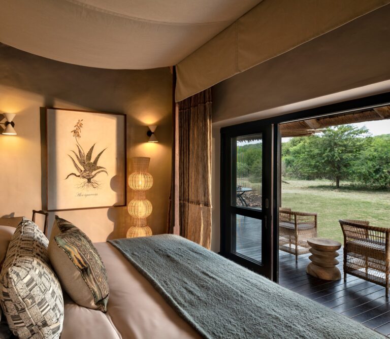 Cozy bedroom with a bed, patterned pillows, and a textured throw, near large glass doors opening to a sunny outdoor seating area.