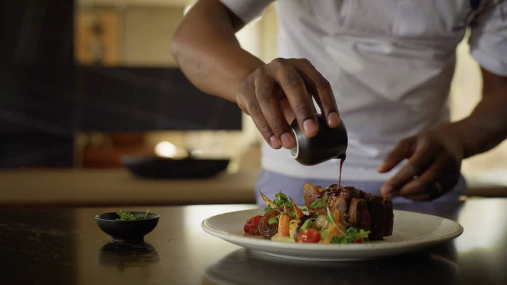 Chef drizzling dark sauce over a plated dish of meat and vegetables, in a kitchen setting.